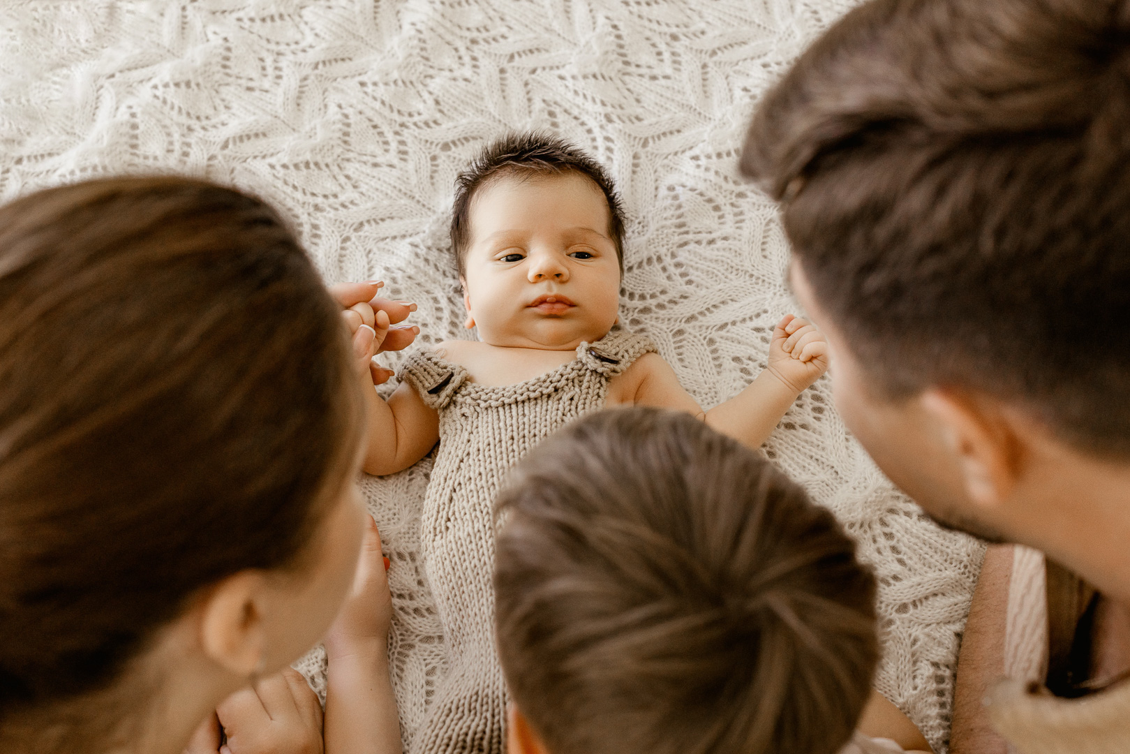 Las Vegas lifestyle newborn photography session — baby in knitted romper lying on cream blanket, surrounded by parents and sibling | Angela Wolf Photography