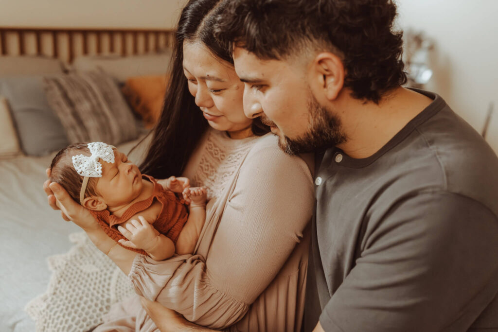 Las Vegas in-home newborn photography — parents lovingly holding baby girl with lace bow during lifestyle session by Angela Wolf Photography.